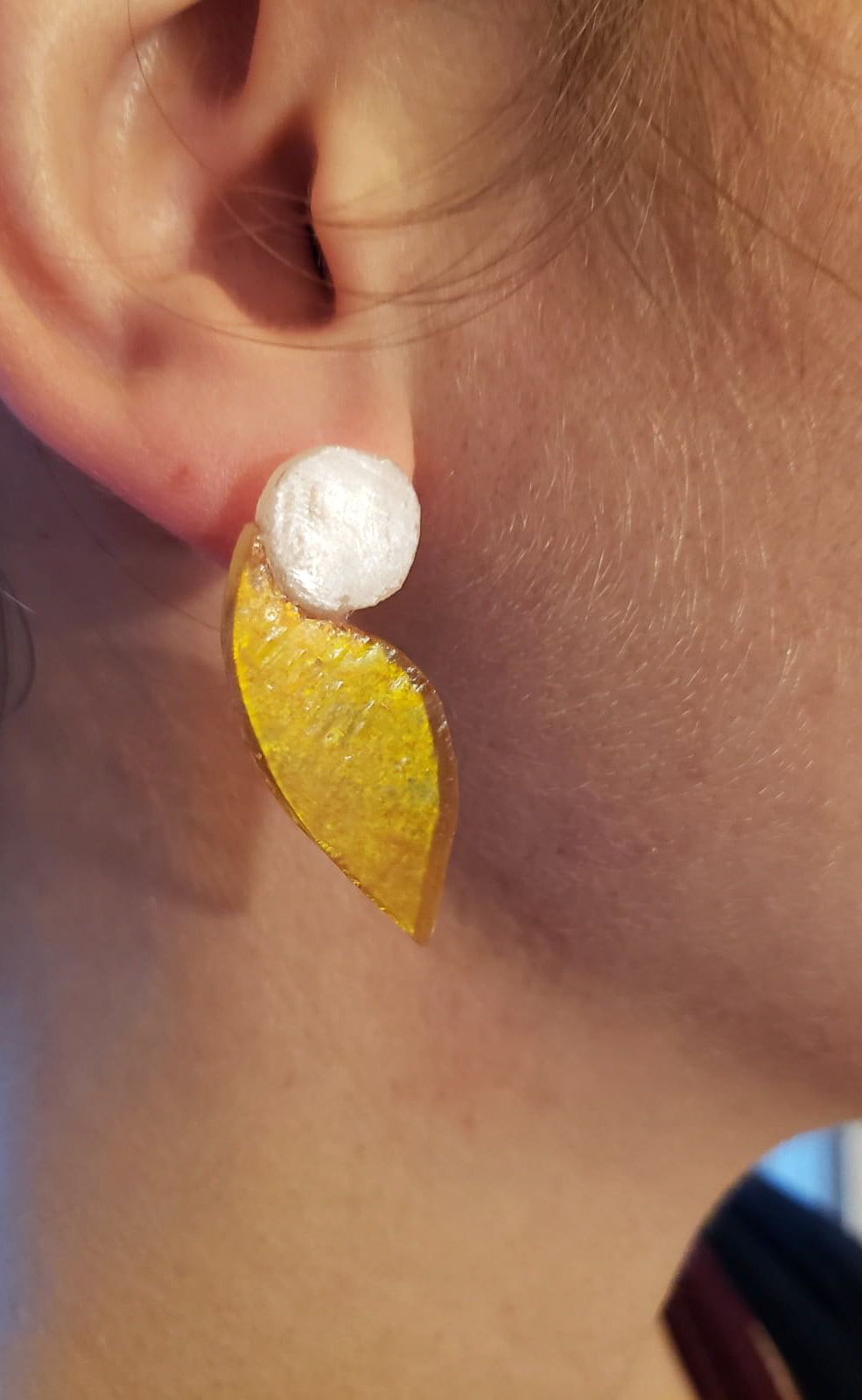 Ces boucles d’oreilles présentent une forme d’aile dans un verre jaune d’or aux reflets irisés, très changeant en fonction de la lumière. Elles sont surmontées d’un disque blanc laiteux. Elles sont agrémentées d’une attache à poussette de laiton doré. 