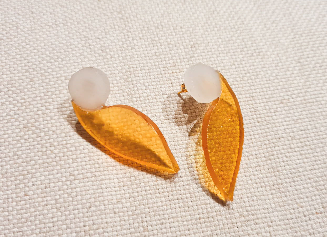 Ces boucles d’oreilles présentent une forme d’aile dans un verre jaune d’or aux reflets irisés, très changeant en fonction de la lumière. Elles sont surmontées d’un disque blanc laiteux. Elles sont agrémentées d’une attache à poussette de laiton doré. 